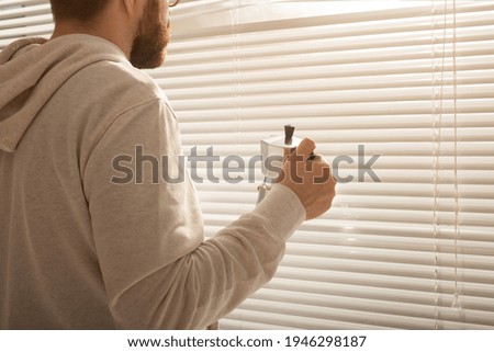 Rear view of young stylish man peeking through hole in window blinds and looking out into street. Concept of enjoying the morning sun and positivity