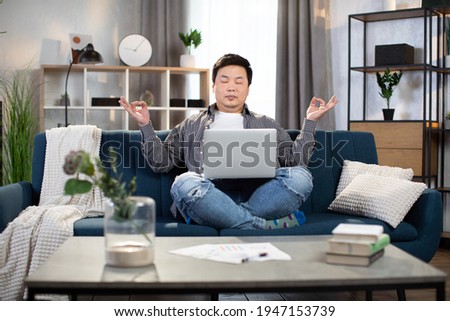 Relaxed asian korean man sitting on couch with laptop on knees and meditating with closed eyes. Tired freelancer taking break during working process at home.