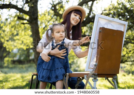 Close up of young charming mother with her little daughter, wearing jeans clothes, painting together on easel outdoors in bright summer garden. Mother\'s Day Concept.