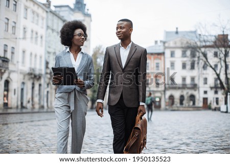 Beautiful woman with clipboard and handsome man with briefcase walking and talking on street. Two african colleagues in stylish formal wear posing outdoors.