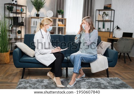 Blond woman doctor in protective medical mask visits her senior patient at home. Therapist is taking notes, while elderly woman is complains of a terrible headache