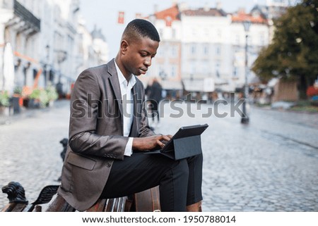 Focused black man in stylish suit sitting on wooden bench outdoors and working on digital tablet. Handsome businessman typing on modern gadget on city street.