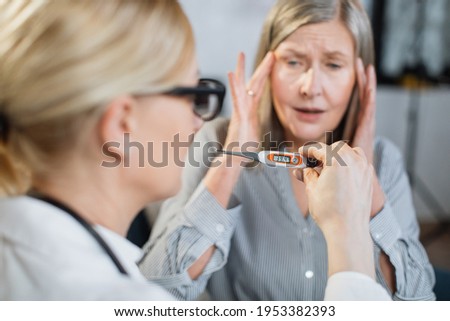 Close up blurred view of female blond doctor holding electronic digital thermometer showing high body temperature and shocked worried retired woman patient scared of her desease. Focus on thermometer.