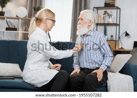 Senior calm bearded man sitting on the couch while caring experienced nice female doctor listening his heartbeat during home visit,front view