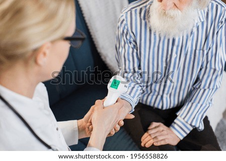 Close up cropped shot of female doctor checking body temperature of senior bearded man patient. Body temperature measurement with infrared wrist non-contact thermometer
