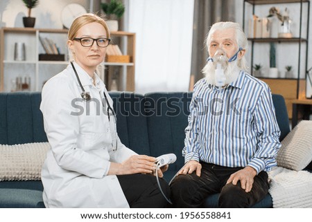 Portrait of senior grey-haired bearded man patient wearing nebulizer inhaler mask, sitting with his smiling professional doctor, looking at camera. Doctor visiting patient at home for inhalation.