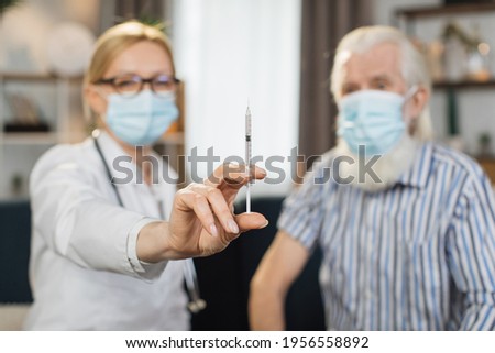 Blurred view of woman doctor and senior man patient, in protective masks, sitting together on the couch at home. Female doctor demonstrating syringe to camera, visiting patient at home.