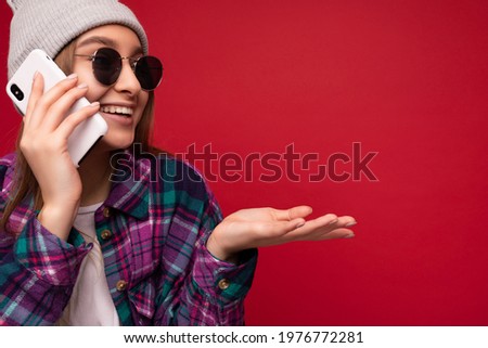 Closeup photo of beautiful emotional positive young blonde woman wearing hipster purple shirt and casual white t-shirt grey hat and sunglasses isolated over red background holding in hand and talking