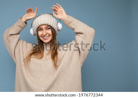 Attractive positive smiling young blonde woman wearing beige winter sweater and hat isolated over blue background wall wearing white wireless bluetooth earphones listening to music having fun and