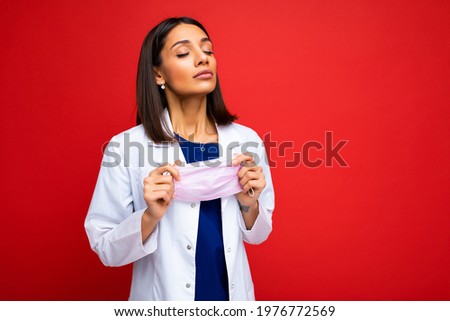 Brunette beautiful Woman taking off virus protective mask on face against coronavirus and white medical coat isolated on red background