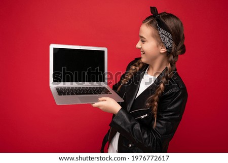 side profile Photo of beautiful happy smiling girl with brunet pigtails holding computer laptop wearing black jacket and bandana isolated over red wall background looking at netbook monitor
