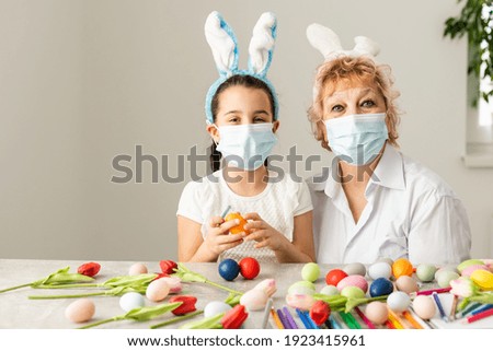 grandmother and granddaughter paint Easter eggs in the kitchen with tassels, phone, tulips and rabbits at home on quarantine with medical mask. Coronavirus, illness, infection, quarantine, flue