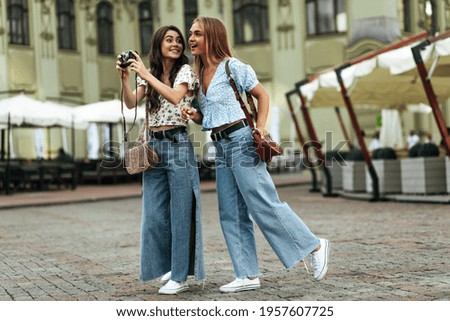 Cool young girlfriends in floral blouses and jeans smile, talk and walk outside. Brunette tanned woman in stylish denim pants holds retro camera.