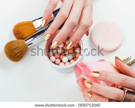 woman hands with golden manicure and many rings holding brushes, makeup artist stuff stylish, pure closeup pink flower rose among cosmetic for makeup