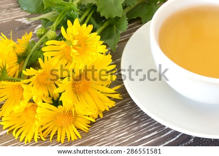Bunch of coltsfoot near cup of herb tea on wooden background