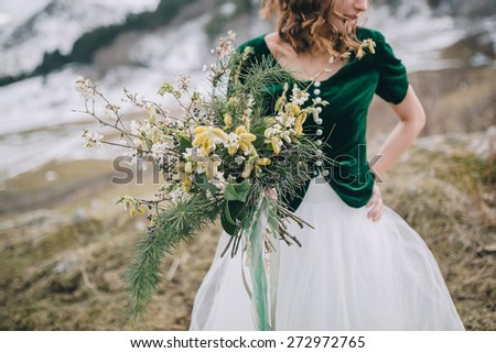 Bride holding wedding bouquet of pine branches and greenery in rustic style on a background of snowy mountains