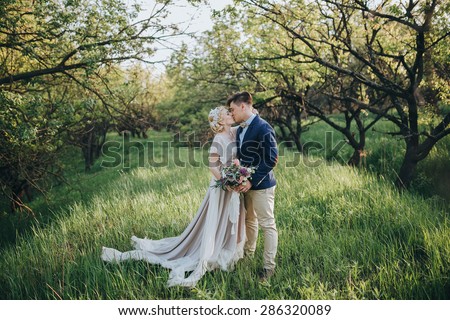 couple in wedding attire with a bouquet of flowers and greenery is in the hands against the backdrop of the garden at sunset, the bride and groom