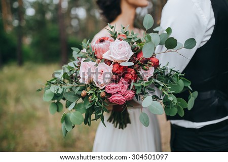 guy in a white shirt and waistcoat and a girl in a white wedding dress with a bouquet of red flowers and greens in hands stand in a clearing in the woods