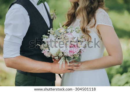 guy in a vest and a girl in a white wedding dress with a bouquet in hands standing in a clearing in the woods