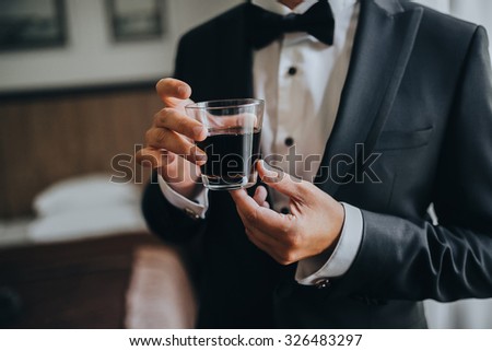 a man in a tuxedo and bow tie, standing and holding a glass of dark liquid