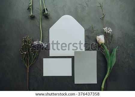 Details. Decor. On a gray textural background is white envelope for invitations, letterhead for the invitation, and nearby is a bouquet of dried flowers, tulips and purple wild flower