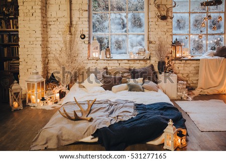 near snowy window is the bed with pillows and blankets, there are a number candles, Christmas decorations and Garlands