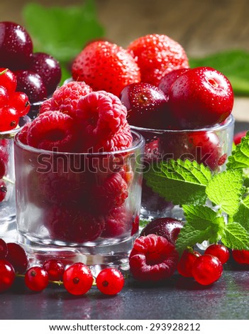 Assorted summer berries: raspberries, red currant, cherry, strawberry with glasses on black background, selective focus