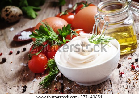 Homemade mayonnaise sauce in a white bowl, jar with olive oil, eggs, salt, spices, mustard, herbs, cherry tomatoes on old wooden background, selective focus