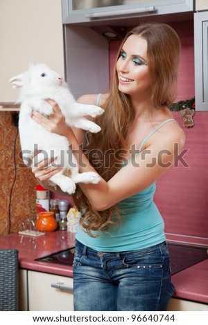 Attractive young woman with white rabbit in the hands smiling and joyful looking out of the camera in the modern kitchen