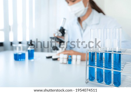 Scientist woman in medical face mask using microscope and test tube with blue liquid while working to research and experiment about coronavirus vaccine in laboratory