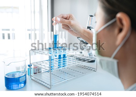Scientist woman in medical face mask is holding test tube with blue liquid and looking reaction of experimental coronavirus vaccine while working with microscope in laboratory