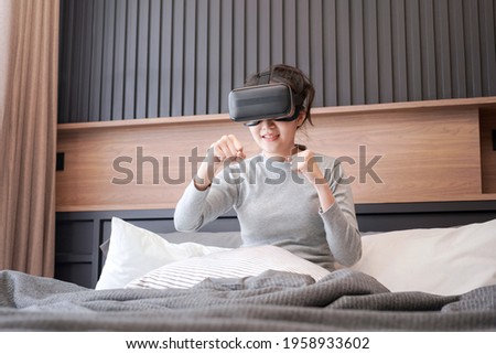 Young asian woman in casual clothes wearing virtual reality glasses to playing fight in boxing game while rest and sitting on the bed in home bedroom