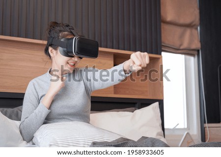Young asian woman in casual clothes wearing virtual reality glasses to playing fight in boxing game while rest and sitting on the bed in home bedroom
