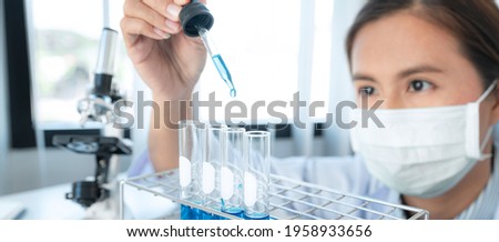 Scientist woman in medical face mask is holding dropper to dropping chemical liquid into test tube and looking reaction of experimental coronavirus vaccine while working in laboratory