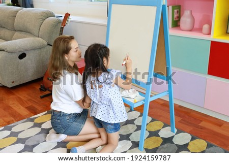 Rear view happy Asian family single mom teaching her little girl how to drawing picture on white board in the living room with happy smiling face (eduction concept)