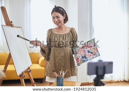 Creative influencer an online streaming painting demonstration. A young female Asian artist holding a paint palette and paint brushes is broadcasting live with a smartphone at home.