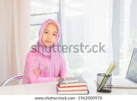 Happy Asian Muslim girl wearing hijab and looking at camera while sitting at the desk with arms crossed, Portrait of Islam kid in the living room.