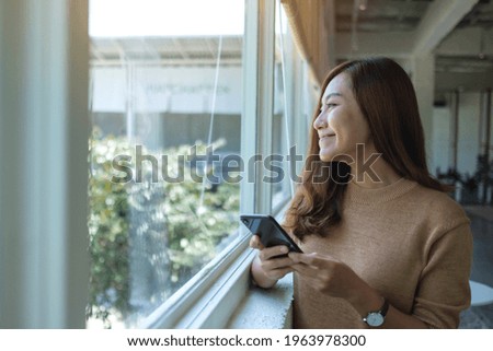 Portrait image of a beautiful young asian woman looking outside the window while holding and using mobile phone