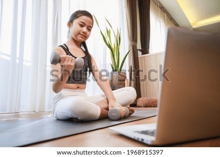 Little Girl doing dumbbell exercise by using online learning with a laptop computer on a yoga mat in the living room at home. New normal concept. Healthy lifestyle. Relaxing at home