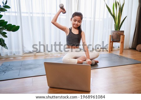 Little Girl doing dumbbell exercise by using online learning with a laptop computer on a yoga mat in the living room at home. New normal concept. Healthy lifestyle. Relaxing at home