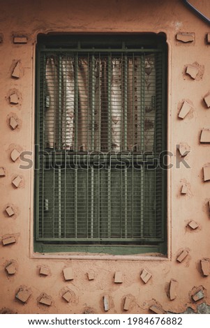 A vertical shot of a soft pink house wall with stone pieces glued around the green bars of a window