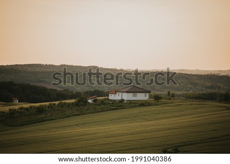 A simple house on a green field under a golden sky with forests and hills in the background