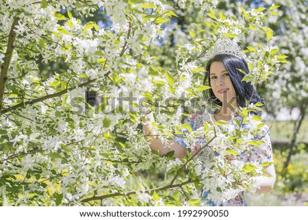 A young beautiful brunette girl in a summer blue dress and with a diadem on her head stands among the flowers of the apple tree  Good spring mood