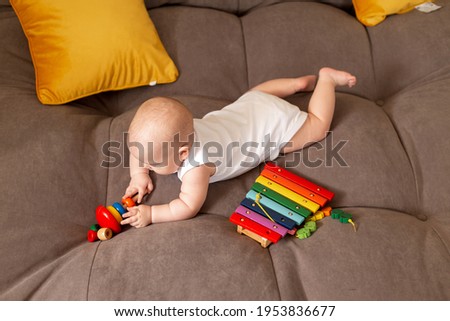 Cute toddler in white bodysuit lies at home on grey sofa with yellow pillows playing with wooden developing toy