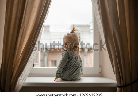 Cute baby blonde girl in vintage dress stands by the window of the house