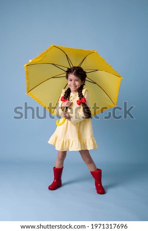 Happy cute little Girl Indian in red rubber boots, cotton dress holds in her hands yellow umbrella on a blue background in the studio. concept Spring, space for text