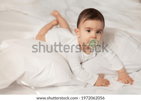 cute little mestizo girl lying in bed with a pacifier in her mouth in a white bodysuit with cotton bedding. High quality photo