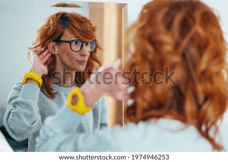 Young redhead woman putting a hearing aid in her ear while looking herself in a mirror