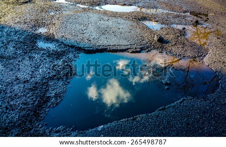Water puddle pothole in concrete cement on road or sidewalk in urban industrial setting. Water reflection of clouds and tree branches. Urban art, street art, abstract art.