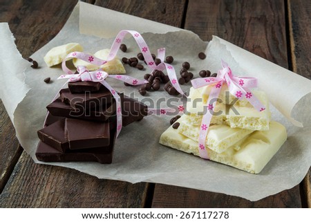 Pieces of white and dark chocolate, tied with a ribbon, chocolate pieces and drops on paper and wooden table. Selective focus